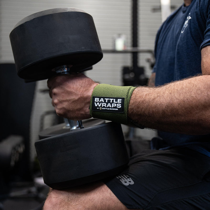 Man wearing Battle Bunker wrist wraps lifting large black dumbbell in gym setting