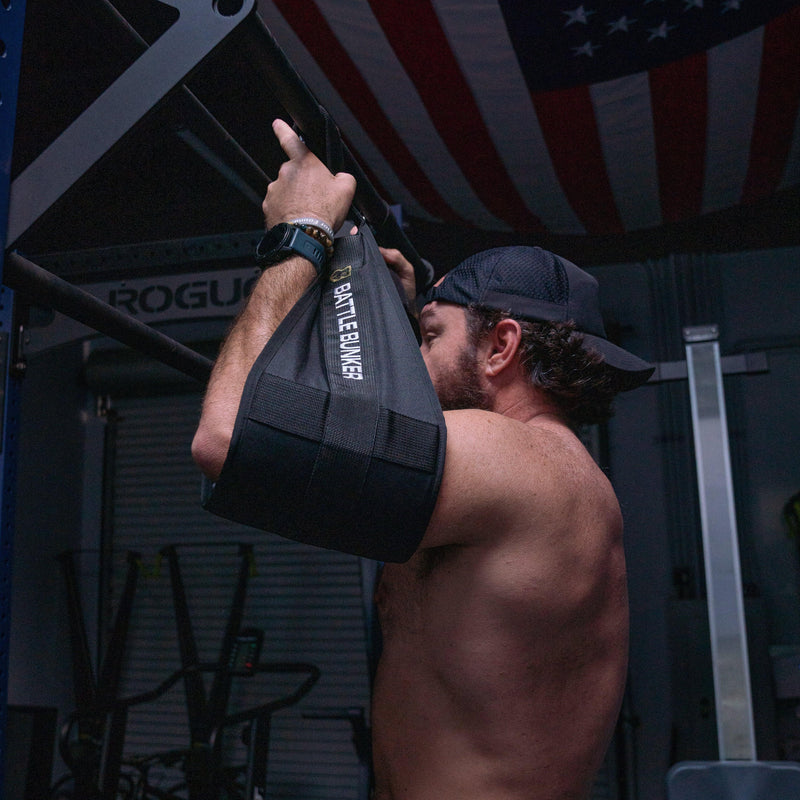 Shirtless man wearing a backwards cap using Battle Bunker arm sling for pull-up in gym
