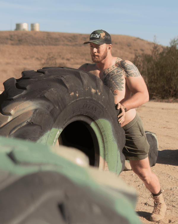 Shirtless man with tattoo flipping large Titan tire outdoors in Battle Bunker workout setting