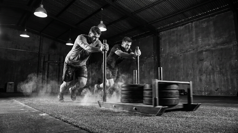Athletes driving a heavily loaded sled push across turf in a gritty functional fitness gym