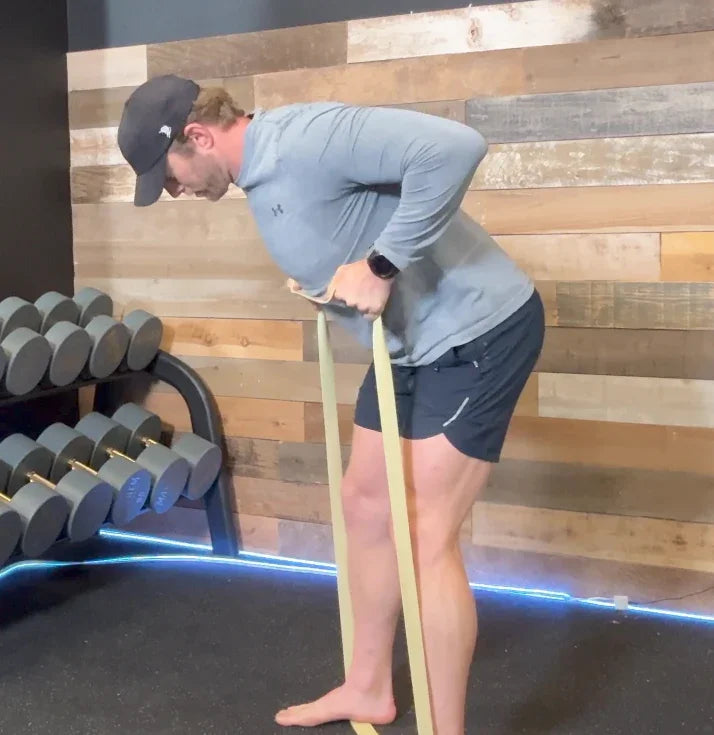 Man in battle bunker gym using resistance bands for bent-over rows near dumbbell rack against wood wall