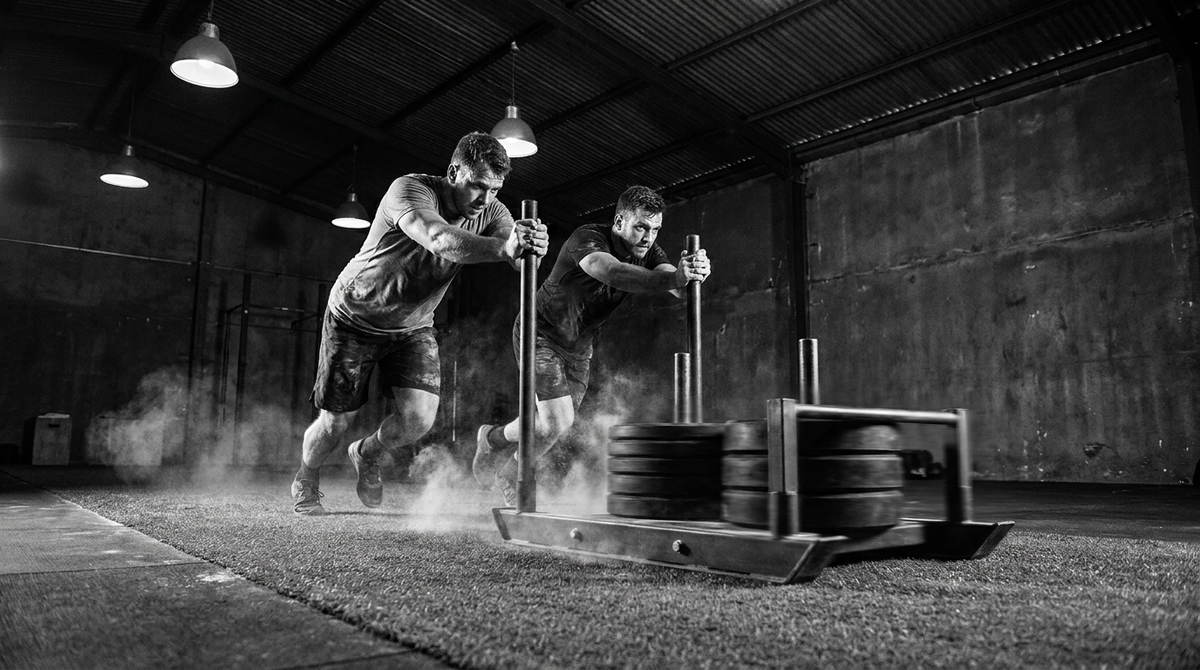 Athletes driving a heavily loaded sled push across turf in a gritty functional fitness gym