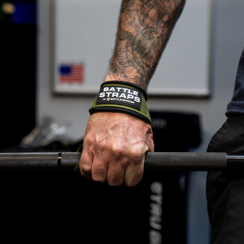 Close-up of tattooed hand gripping barbell wearing Battle Bunker green Battle Straps wrist support