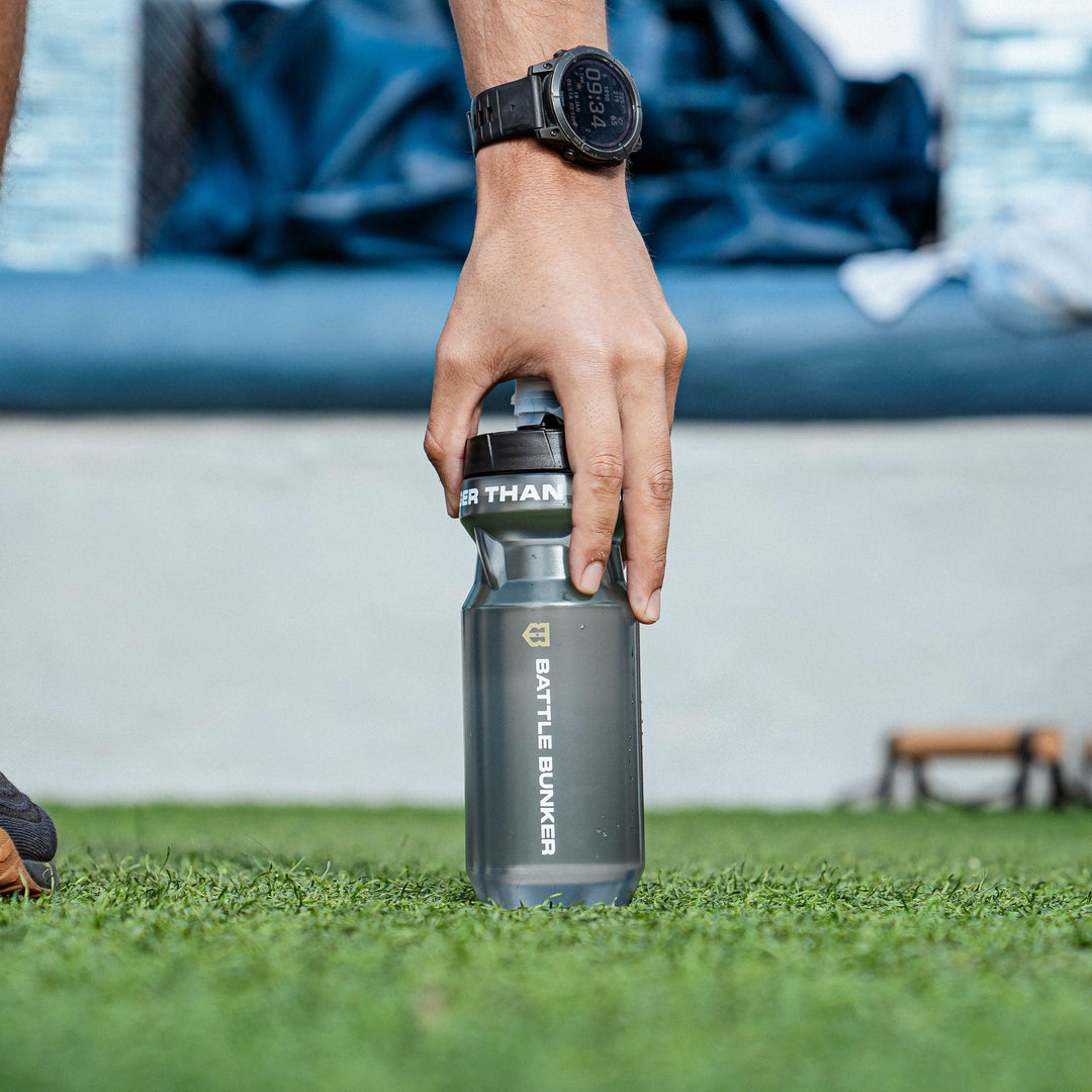 Hand holding a gray Battle Bunker water bottle on grass with a digital watch on wrist