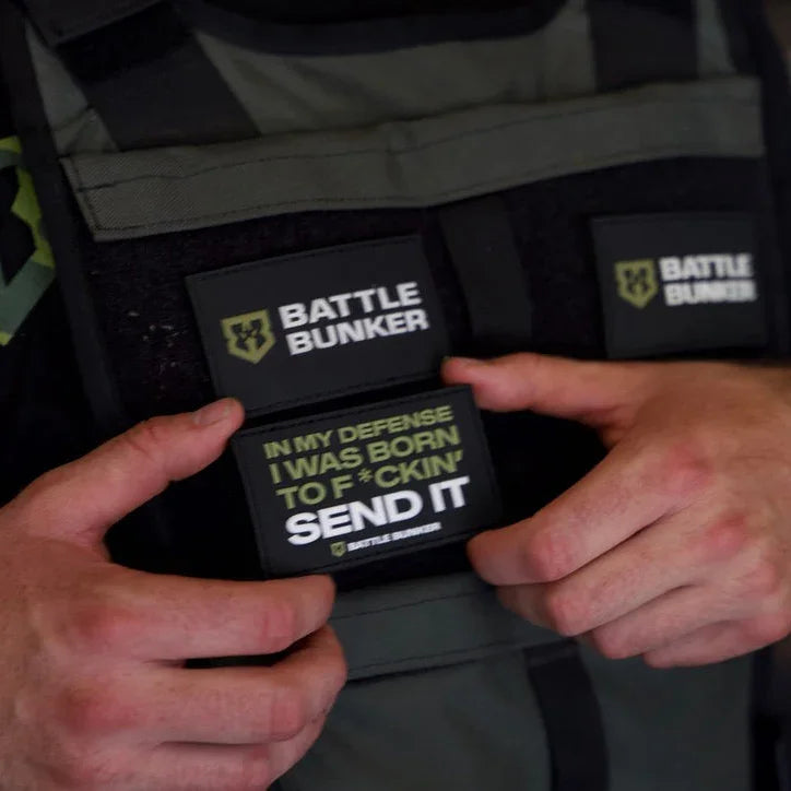 Hands holding a Battle Bunker patch on tactical vest with 'In my defense I was born to f*ckin send it' slogan patch