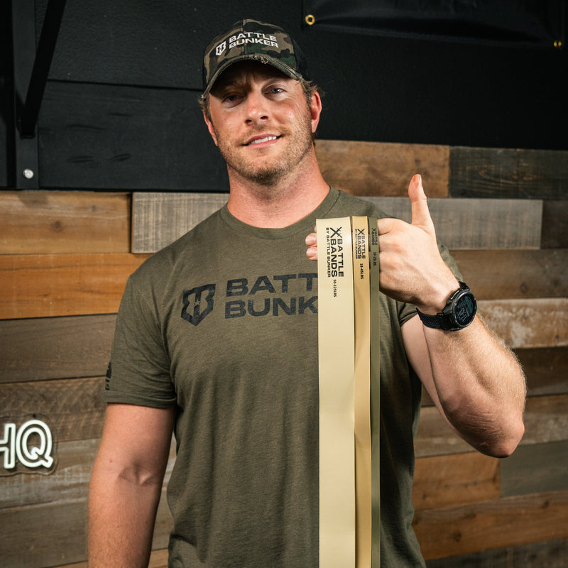 Austen Alexander wearing an olive green Batte Bunker t-shirt and a black cap gives a thumbs-up while holding a Batt Bunk resistance band. The background features a rustic wooden wall.