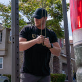 Man in black Battle Bunker t-shirt adjusting resistance bands outdoors near home gym rack