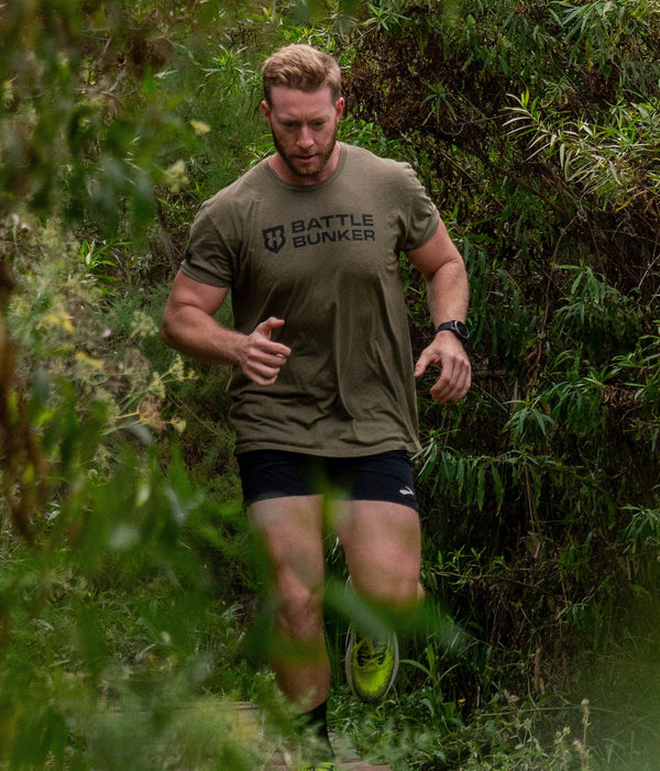 Man running on forest trail wearing olive Battle Bunker t-shirt and black shorts