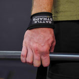 Hand gripping a barbell with Battle Bunker wrist straps in a gym setting