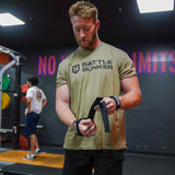 Man in olive Battle Bunker t-shirt wrapping wrist straps in gym with weightlifting equipment