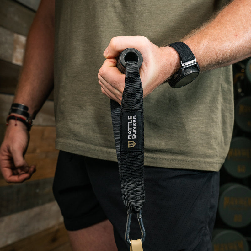 A close-up of a person's hand holding a black resistance band handle with a black webbing strap featuring the 'BATTLE BUNKER' brand logo. The person is wearing a green t-shirt and black athletic shorts in a gym setting with wooden walls and weight plates in the background.