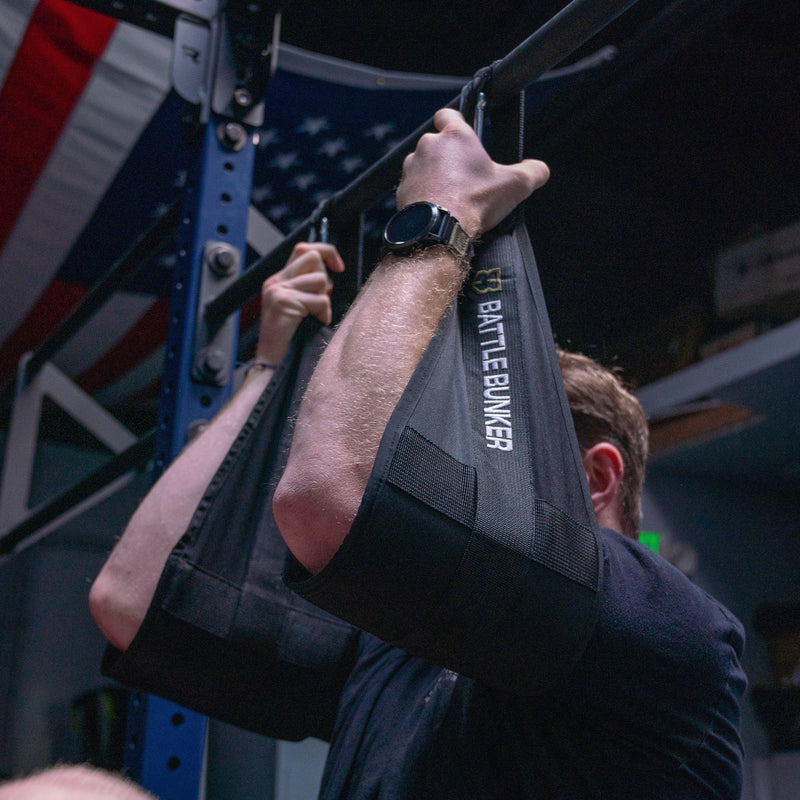 Person doing pull-up with black Battle Bunker arm slings in a gym with American flag background