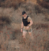 Man wearing black tactical battle bunker vest and cap walking through dry brush in outdoor setting