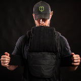 Man wearing Battle Bunker cap holding open black tactical vest with Velcro panels against black background