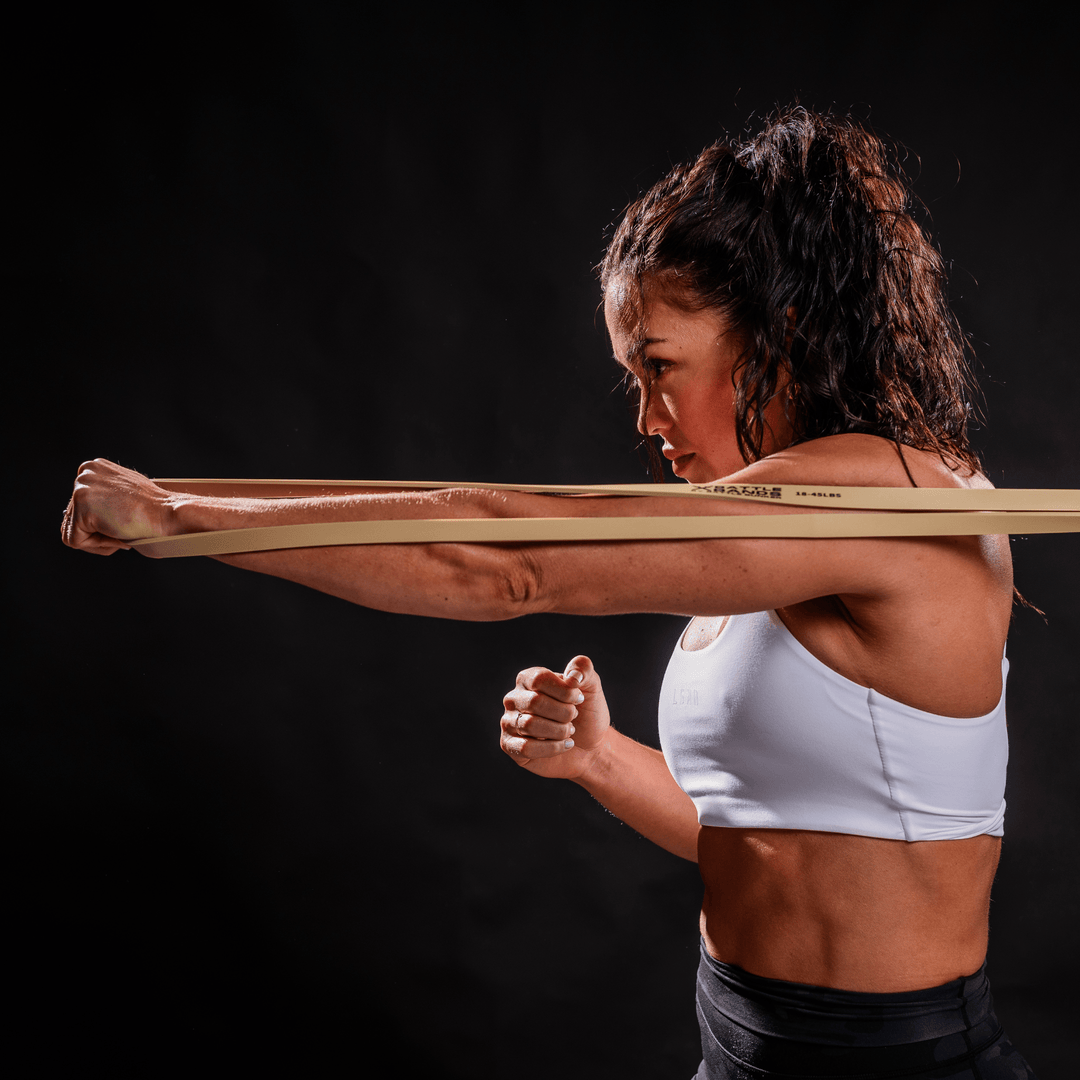 Woman in white sports bra using Battle Bunker resistance band for arm workout against black background