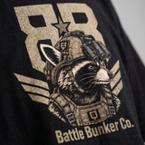 Close-up of a black graphic t-shirt featuring a distressed beige print of a raccoon in tactical gear with a star helmet, the number 88, and the text 'Battle Bunker Co.'.