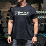 Man wearing black Battle Bunker t-shirt in gym with dumbbells and weights in background