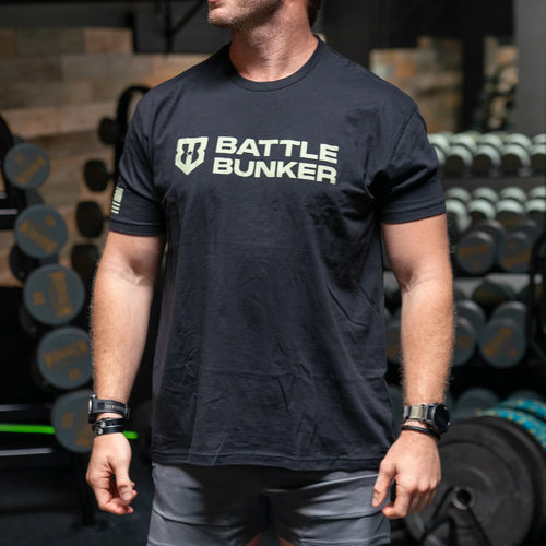 Man wearing black Battle Bunker t-shirt in gym with dumbbells and weights in background