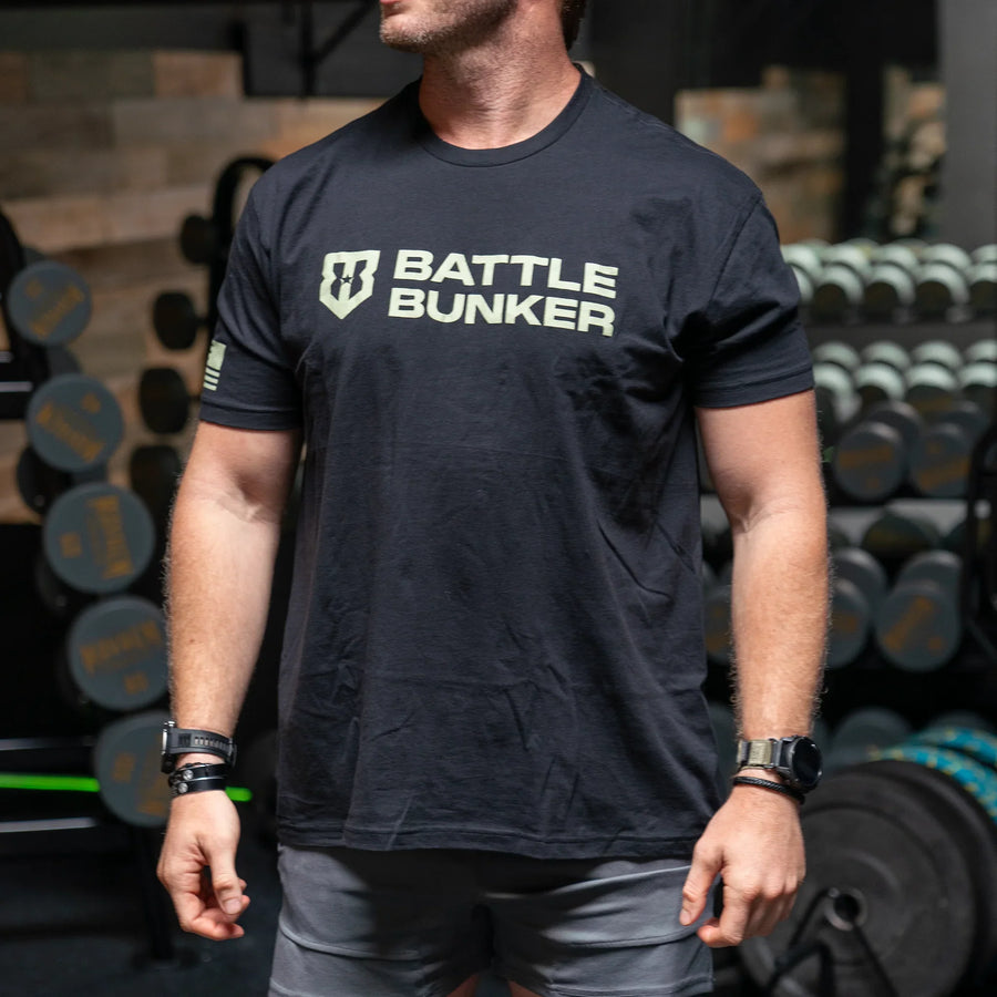 Man wearing black Battle Bunker t-shirt in gym with dumbbells and weights in background