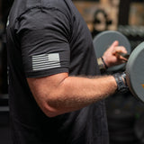Close-up of muscular arm wearing black Battle Bunker t-shirt with American flag lifting dumbbell