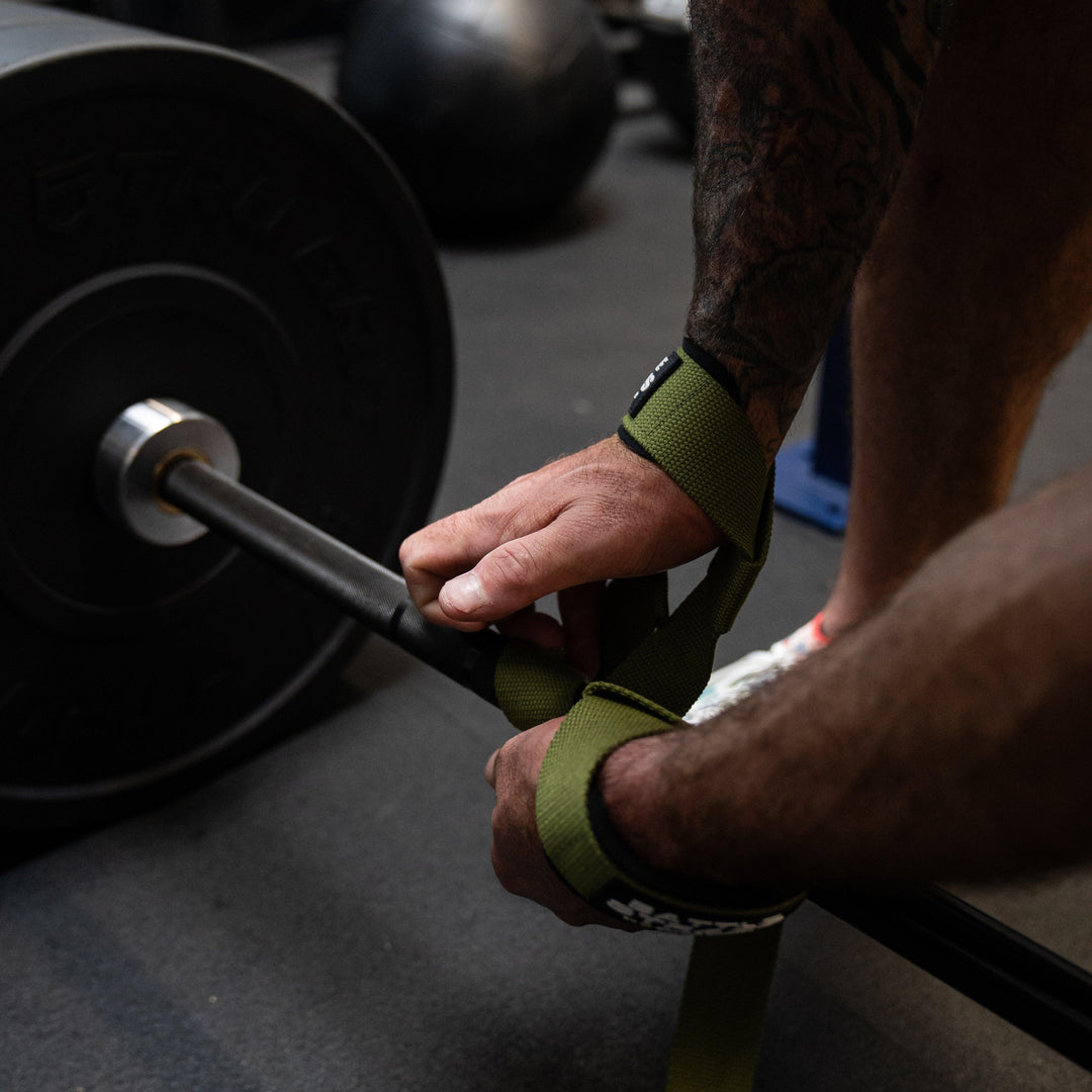 Close-up of tattooed hands attaching green Battle Bunker lifting straps to barbell in gym setting