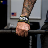Close-up of tattooed hand wearing green Battle Bunker battle straps gripping a textured barbell in gym